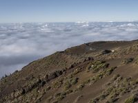 an empty road is descending above the clouds below a hill near a town on a hilltop